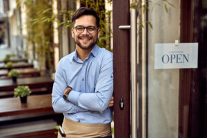 Happy waiter with arms crossed standing next to open sign at the cafe's entrance and looking at the camera